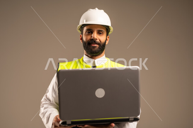 الإعمار بسواعد أبناء الوطن، استخدام الأجهزة التقنية الحديثة في الهندسة والانشاءات المعمارية، بورتريه مقرب لمهندس عربي خليجي سعودي يرتدي الثوب التقليدي والسترة الخاصة وخوذة الحماية يحمل الحاسوب المحمول ينظر إلى الكاميرا يقوم بمتابعة الأعمال، خلفية بيج