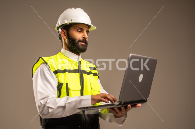 استخدام الأجهزة التقنية الحديثة في الهندسة والانشاءات المعمارية، بورتريه مقرب لمهندس عربي خليجي سعودي يرتدي الثوب التقليدي والسترة الخاصة وخوذة الحماية يحمل الحاسوب المحمول يقوم بمتابعة الأعمال، الإعمار بسواعد أبناء الوطن، خلفية بيج