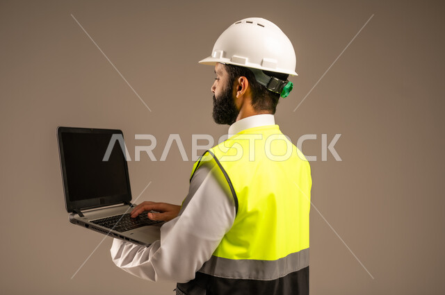 استخدام الأجهزة التقنية الحديثة في الهندسة والانشاءات المعمارية، بورتريه مقرب من الجانب لمهندس عربي خليجي سعودي يرتدي الثوب التقليدي والسترة الخاصة وخوذة الحماية يحمل الحاسوب المحمول يقوم بمتابعة الأعمال، الإعمار بسواعد أبناء الوطن، خلفية بيج