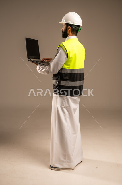 Using modern technical devices in engineering and architectural construction, side portrait of a Saudi Arabian Gulf engineer wearing traditional dress, special jacket and protective helmet, holding a laptop computer following up on work, construction by the hands of the sons of the homeland, full body, beige background