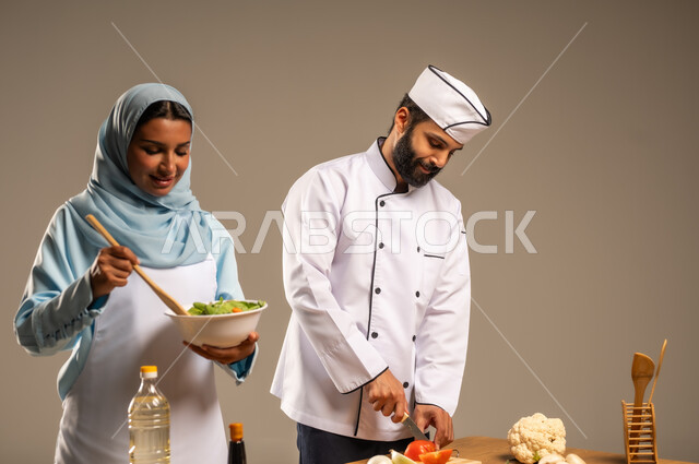 المشاركة في تحضير الوصفات الشهية, الاستمتاع بالعمل الجماعي في مهنة الطبخ، بورتريه مقرب لرجل وامرأة عربيين خليجيين سعوديين يقفان خلف الطاولة الخشبية ويتشاركان في تجهيز الطعام، إعداد السلطات والخضروات والأطباق الصحية، ارتداء زي الشيف والمئزر الأبيض، خلفية رمادية