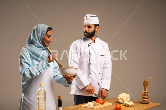 المشاركة في تحضير الوصفات الشهية, الاستمتاع بالعمل الجماعي في مهنة الطبخ، بورتريه مقرب لرجل وامرأة عربيين خليجيين سعوديين يقفان خلف الطاولة الخشبية ويتشاركان في تجهيز الطعام، إعداد السلطات والخضروات والأطباق الصحية، ارتداء زي الشيف والمئزر الأبيض، خلفية رمادية