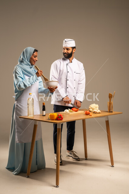 الاستمتاع بالعمل الجماعي في مهنة الطبخ، المشاركة في تحضير الوصفات الشهية, بورتريه لرجل وامرأة عربيين خليجيين سعوديين يقفان خلف الطاولة الخشبية ويتشاركان في تجهيز الطعام، إعداد الأطباق الصحية، ارتداء زي الشيف والمئزر الأبيض، الجسم بالطول الكامل، خلفية رمادية