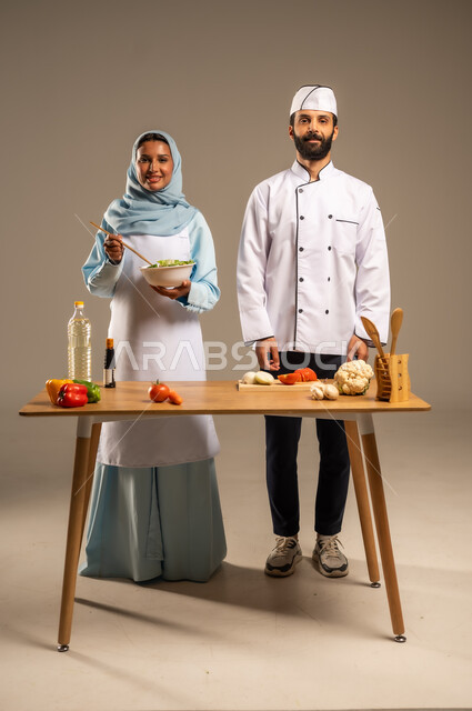 الاستمتاع بالعمل الجماعي في مهنة الطبخ، المشاركة في تحضير الوصفات الشهية, بورتريه لرجل وامرأة عربيين خليجيين سعوديين يقفان خلف الطاولة الخشبية ويتشاركان في تجهيز الطعام، إعداد الأطباق الصحية، ارتداء زي الشيف والمئزر الأبيض، الجسم بالطول الكامل، خلفية رمادية