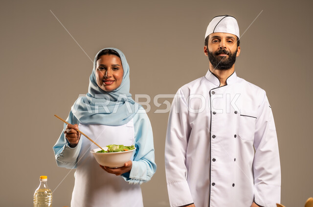 المشاركة في تحضير الوصفات الشهية, الاستمتاع بالعمل الجماعي في مهنة الطبخ، بورتريه مقرب لرجل وامرأة عربيين خليجيين سعوديين يقفان خلف الطاولة الخشبية ويتشاركان في تجهيز الطعام، إعداد السلطات والخضروات والأطباق الصحية، ارتداء زي الشيف والمئزر الأبيض، خلفية رمادية