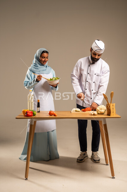 الاستمتاع بالعمل الجماعي في مهنة الطبخ، المشاركة في تحضير الوصفات الشهية, بورتريه لرجل وامرأة عربيين خليجيين سعوديين يقفان خلف الطاولة الخشبية ويتشاركان في تجهيز الطعام، إعداد الأطباق الصحية، ارتداء زي الشيف والمئزر الأبيض، الجسم بالطول الكامل، خلفية رمادية