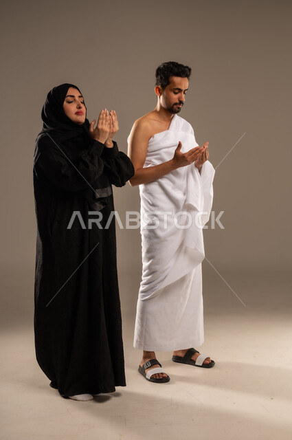 Maintaining prayers, supplications and asking forgiveness from Allah Almighty, answering Allah's call to perform Hajj, portrait of a Saudi Arabian Gulf couple wearing Ihram clothes, supplicating with humility and supplication, the fifth pillar of Islam, full body image, beige background