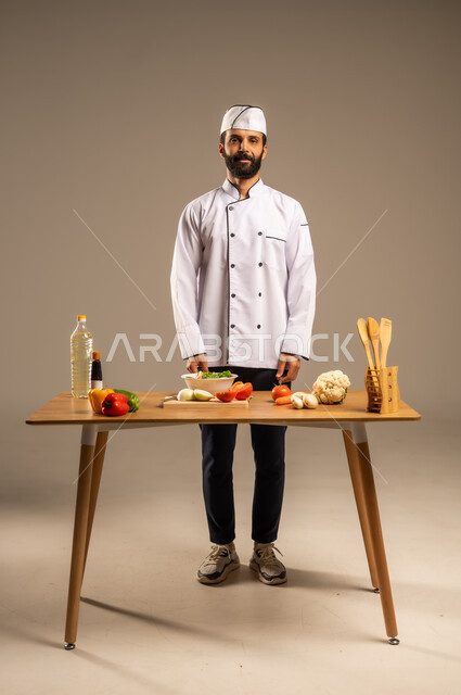 Professional chef, skills of presenting and decorating food in distinctive and innovative ways, enjoyment in preparing delicious recipes, close-up portrait of a young Saudi Arabian Gulf man wearing a special chef's uniform with a white hat and preparing healthy salad dishes, full-length body, gray background