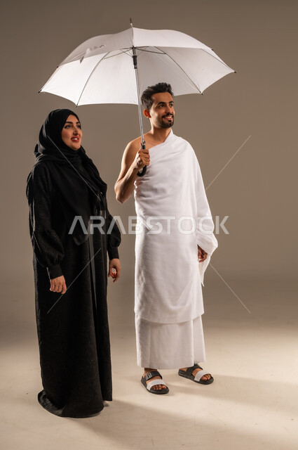 استخدام المظلة الشمسية للحماية والتظلل،  الاستعداد لأداء مناسك الحج والعمرة، بورتريه لزوجين عربيين خليجيين سعوديين يرتديان ثياب الاحرام ينظران الى مكان ما بتعابير الفرح والسرور، مفهوم العبادة والتقرب الى الله تعالى، زيارة بيت الله الحرام، صورة الجسم بالكامل، خلفية بيج