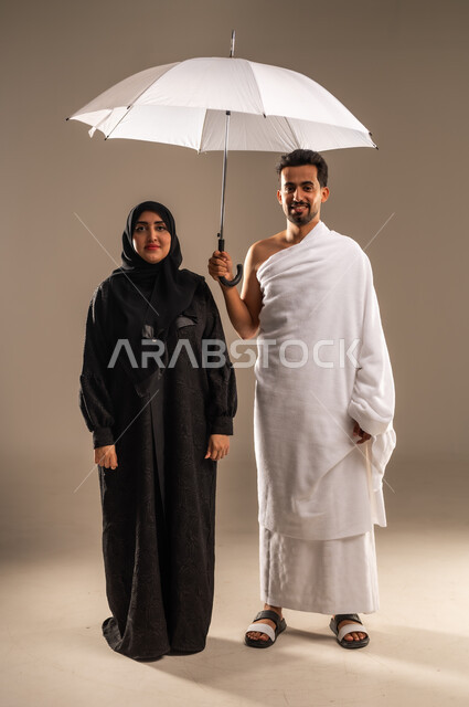 استخدام المظلة الشمسية للحماية والتظلل، بورتريه لزوجين عربيين خليجيين سعوديين يرتديان ثياب الإحرام لأداء مناسك الحج والعمرة، النظر الى الكاميرا بتعابير الفرح والسرور، مفهوم العبادة والتقرب الى الله تعالى، زيارة بيت الله الحرام، صورة الجسم بالكامل، خلفية بيج