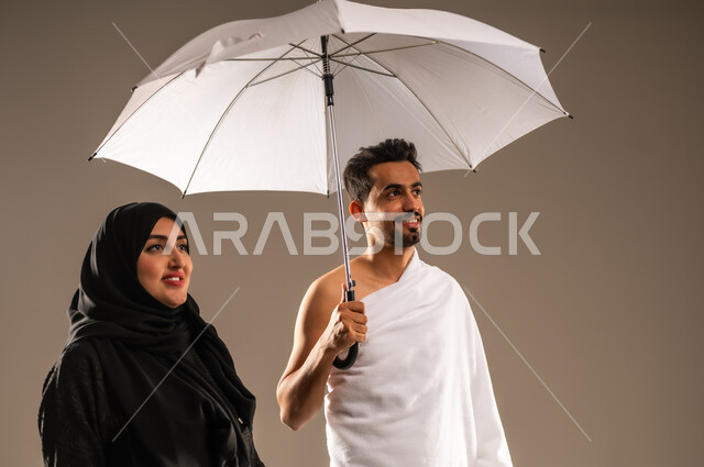 مفهوم العبادة والتقرب الى الله تعالى، استخدام المظلة الشمسية للحماية والتظلل، بورتريه لزوجين عربيين خليجيين سعوديين يرتديان ثياب الإحرام لأداء مناسك الحج والعمرة، ايماءات الفرح والسرور لزيارة بيت الله الحرام، خلفية بيج