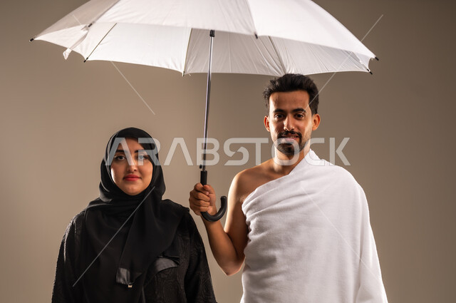 The concept of worship and getting closer to God Almighty, using a sun umbrella for protection and shade, a portrait of a Saudi Arabian Gulf couple wearing Ihram clothes to perform Hajj and Umrah rituals, gestures of joy and happiness for visiting the Holy House of God, beige background
