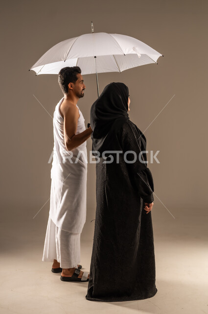 استخدام المظلة الشمسية للحماية والتظلل،  الاستعداد لأداء مناسك الحج والعمرة، بورتريه مقرب من الجانب لزوجين عربيين خليجيين سعوديين يرتديان ثياب الاحرام ينظران الى مكان ما بتعابير الفرح والسرور، مفهوم العبادة والتقرب الى الله تعالى، زيارة بيت الله الحرام، صورة الجسم بالكامل، خلفية بيج