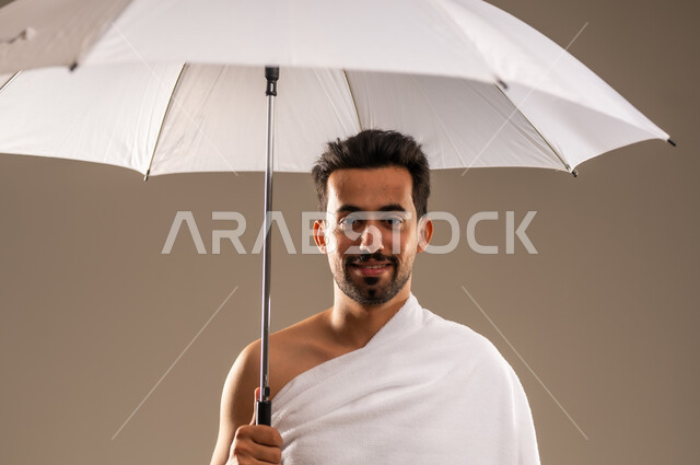 العبادة والتقرب الى الله بالأعمال الصالحة، أداء مناسك فريضة الحج و العمرة، زيارة بيت الله الحرام في مكة المكرمة، ايماءات السعادة و الطمأنينة، بورتريه مقرب لرجل مسلم عربي خليجي سعودي مبتسم يرتدي ثياب الاحرام يحمل بيده مظلة شمسية بيضاء للحماية والتظلل، خلفية بيج