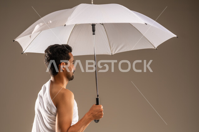 العبادة والتقرب الى الله بالأعمال الصالحة، أداء مناسك فريضة الحج و العمرة، زيارة بيت الله الحرام في مكة المكرمة، بورتريه مقرب من الخلف لرجل مسلم عربي خليجي سعودي مبتسم يرتدي ثياب الاحرام يحمل بيده مظلة شمسية بيضاء للحماية والتظلل، خلفية بيج