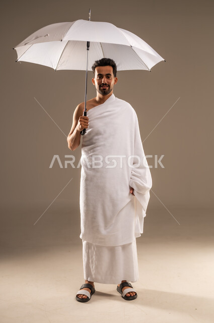 The concept of worship and getting closer to God, performing the rituals of Hajj and Umrah, visiting the Holy House of God in Mecca, looking at the camera with expressions of happiness and reassurance, close-up portrait of a smiling Saudi Arabian Gulf Arab Muslim man wearing Ihram clothes holding a sun umbrella for protection and shade, full body length, beige background