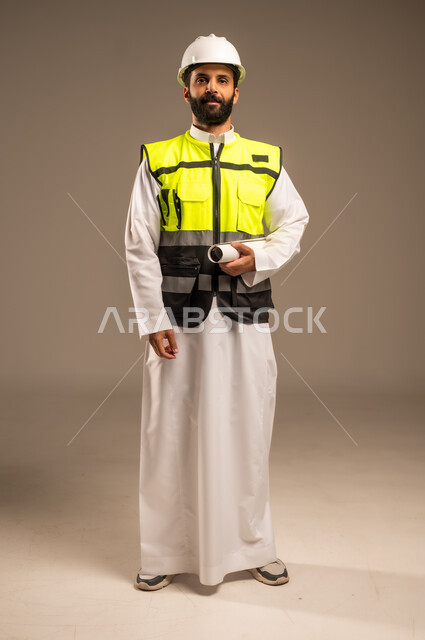 الإعمار بسواعد أبناء الوطن، مفهوم الهندسة والانشاءات المعمارية، بورتريه لمهندس عربي خليجي سعودي يرتدي الثوب التقليدي والسترة الخاصة وخوذة الحماية يحمل مخطط الإعمار ينظر إلى الكاميرا يقوم بمتابعة الأعمال، مهن ووظائف سعودية للشباب، الجسم بالطول الكامل، خلفية بيج