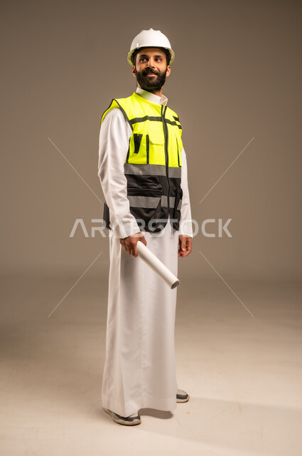 الإعمار بسواعد أبناء الوطن، مفهوم الهندسة والانشاءات المعمارية، بورتريه لمهندس عربي خليجي سعودي يرتدي الثوب التقليدي والسترة الخاصة وخوذة الحماية يحمل مخطط الإعمار ينظر إلى شيء ما يقوم بمتابعة الأعمال، مهن ووظائف سعودية للشباب، الجسم بالطول الكامل، خلفية بيج