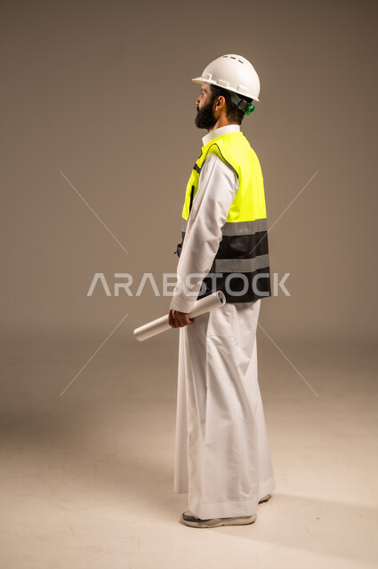 Concept of engineering and architectural construction, side portrait of a Saudi Arabian Gulf engineer wearing traditional dress, special jacket and protective helmet holding construction plan and following up on work, Saudi professions and jobs for young people, construction by the hands of the sons of the homeland, full body, beige background
