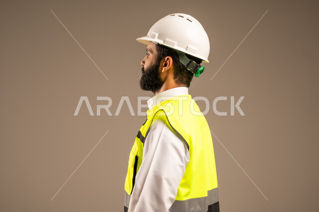 Concept of engineering and architectural construction, close-up portrait from the side of a Saudi Arabian Gulf engineer wearing traditional dress, special jacket and protective helmet looking at something following up on work, Saudi professions and jobs for young people, construction by the hands of the sons of the homeland, beige background