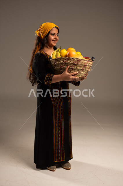 منتجات ومحاصيل زراعية وطنية، ذكرى تأسيس الدولة السعودية 22 فبراير، يوم بدينا 1727م، ارتداء أزياء المنطقة الجنوبية، بورتريه مقرب من الجانب لفتاة عربية خليجية سعودية ترتدي الشيلة والسدرة تحمل بيدها سلة مصنوعة من القش مليئة بالفواكه الطازجة، الجسم الكامل بالطول ، خلفية بيج