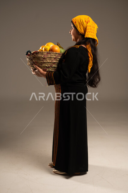 منتجات ومحاصيل زراعية وطنية، ذكرى تأسيس الدولة السعودية 22 فبراير، يوم بدينا 1727م، ارتداء أزياء المنطقة الجنوبية، بورتريه مقرب من الجانب لفتاة عربية خليجية سعودية ترتدي الشيلة والسدرة تحمل بيدها سلة مصنوعة من القش مليئة بالفواكه الطازجة، الجسم الكامل بالطول ، خلفية بيج
