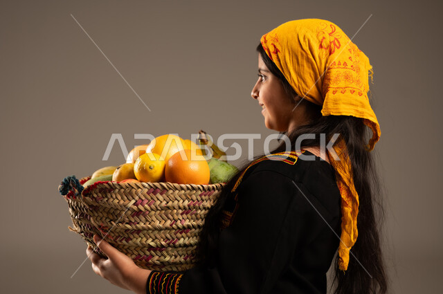 منتجات ومحاصيل زراعية وطنية، ذكرى تأسيس الدولة السعودية 22 فبراير، يوم بدينا 1727م، ارتداء أزياء المنطقة الجنوبية، بورتريه مقرب من الجانب لفتاة عربية خليجية سعودية ترتدي الشيلة والسدرة تحمل بيدها سلة مصنوعة من القش مليئة بالفواكه الطازجة، خلفية بيج