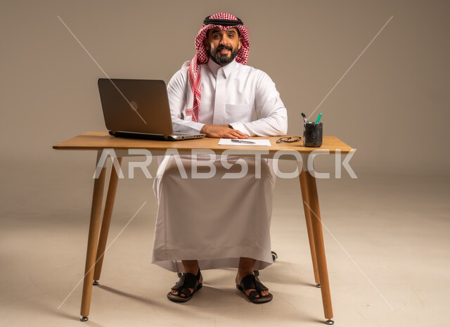 Integrating work with technology and modern technology, office administrative professions and jobs for young people, the concept of managing and organizing business affairs through technical devices, a portrait of a Saudi Arabian Gulf man wearing a traditional thobe and a shemagh sitting behind a desk with a laptop in front of him and a number of files, gray background