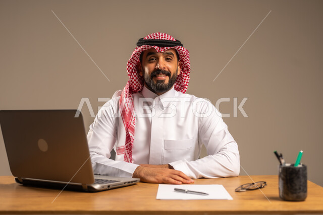 دمج العمل مع التقنية والتكنولوجيا الحديثة، مهن ووظائف إدارية مكتبية للشباب, مفهوم إدارة شؤون الأعمال وتنظيمها عبر الأجهزة التقنية، بورتريه لرجل عربي خليجي سعودي يرتدي الثوب التقليدي والشماغ يجلس خلف المكتب وأمامه الحاسوب المحمول وعدد من الملفات، خلفية رمادية