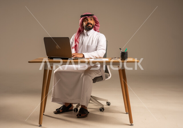 دمج العمل مع التقنية والتكنولوجيا الحديثة، مهن ووظائف إدارية مكتبية للشباب, مفهوم إدارة شؤون الأعمال وتنظيمها عبر الأجهزة التقنية، بورتريه لرجل عربي خليجي سعودي يرتدي الثوب التقليدي والشماغ يجلس خلف المكتب وأمامه الحاسوب المحمول وعدد من الملفات، خلفية رمادية