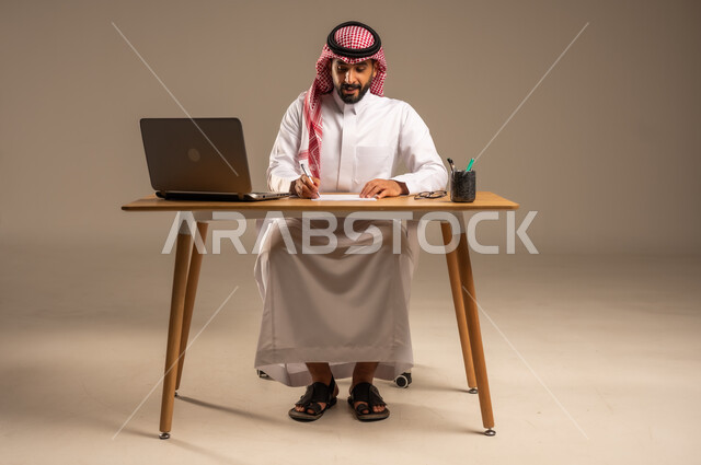 دمج العمل مع التقنية والتكنولوجيا الحديثة، مهن ووظائف إدارية مكتبية للشباب, مفهوم إدارة شؤون الأعمال وتنظيمها عبر الأجهزة التقنية، بورتريه لرجل عربي خليجي سعودي يرتدي الثوب التقليدي والشماغ يجلس خلف المكتب وأمامه الحاسوب المحمول وعدد من الملفات، خلفية رمادية