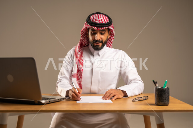 دمج العمل مع التقنية والتكنولوجيا الحديثة، مهن ووظائف إدارية مكتبية للشباب, مفهوم إدارة شؤون الأعمال وتنظيمها عبر الأجهزة التقنية، بورتريه لرجل عربي خليجي سعودي يرتدي الثوب التقليدي والشماغ يجلس خلف المكتب وأمامه الحاسوب المحمول وعدد من الملفات، خلفية رمادية