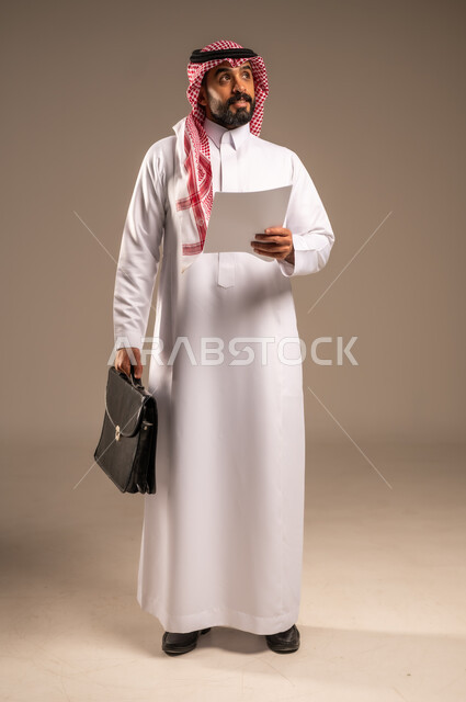 التخطيط للعمل ومراجعة نتائج الاجتماع، إعداد التقارير وتحديد الأهداف اليومية، بورتريه لرجل عربي خليجي سعودي يرتدي الثوب التقليدي والشماغ يحمل بيده مجموعة أوراق وحقيبة يد ينظر إلى شيء ما، تدوين المهام والملاحظات ومراجعتها، الجسم بالطول الكامل، خلفيه بيج