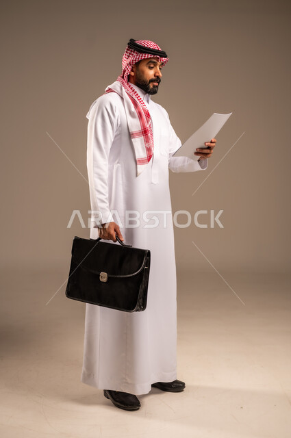 Writing down tasks and notes on paper and reviewing them, side portrait of a Saudi Arabian Gulf man wearing traditional thobe and shemagh holding a bunch of papers and a handbag, planning work and reviewing meeting results, preparing reports and setting daily goals, full body, beige background