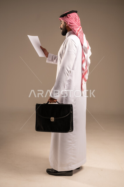 تدوين المهام والملاحظات على الورق ومراجعتها، بورتريه من الجانب لرجل عربي خليجي سعودي يرتدي الثوب التقليدي والشماغ يحمل بيده مجموعة أوراق وحقيبة يد، التخطيط للعمل ومراجعة نتائج الاجتماع، إعداد التقارير وتحديد الأهداف اليومية، الجسم بالطول الكامل، خلفيه بيج