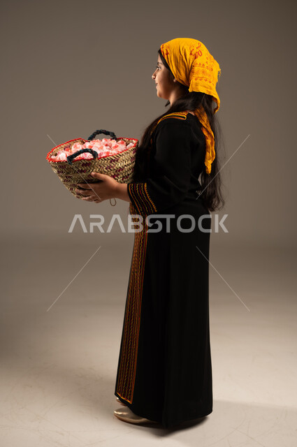 الزهور والورود العطرية، يوم بدينا1727م، بورتريه مقرب من الخلف لفتاة عربية خليجية سعودية مبتسمة ترتدي الشيلة والسدره تحمل بيدها سلة قش ممتلئة بالورد الجوري الطبيعي، ارتداء أزياء المنطقة الجنوبية، ذكرى تاسيس الدولة السعودية 22فبراير، الجسم الكامل بالطول، خلفية بيج 