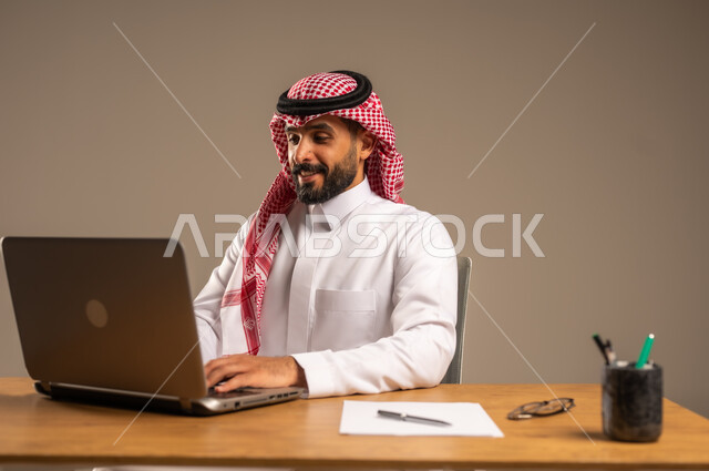 مفهوم إدارة شؤون الأعمال وتنظيمها عبر الأجهزة التقنية، دمج العمل مع التقنية والتكنولوجيا الحديثة، مهن ووظائف إدارية مكتبية للشباب, بورتريه لرجل عربي خليجي سعودي يرتدي الثوب التقليدي والشماغ يجلس خلف المكتب وأمامه الحاسوب المحمول وعدد من الملفات، خلفية رمادية