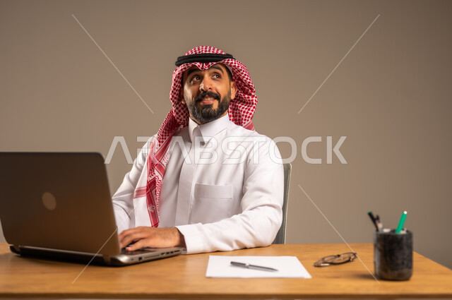 مفهوم إدارة شؤون الأعمال وتنظيمها عبر الأجهزة التقنية، دمج العمل مع التقنية والتكنولوجيا الحديثة، مهن ووظائف إدارية مكتبية للشباب, بورتريه لرجل عربي خليجي سعودي يرتدي الثوب التقليدي والشماغ يجلس خلف المكتب وأمامه الحاسوب المحمول وعدد من الملفات، خلفية رمادية