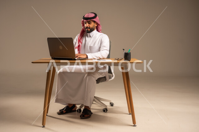 مفهوم إدارة شؤون الأعمال وتنظيمها عبر الأجهزة التقنية، دمج العمل مع التقنية والتكنولوجيا الحديثة، مهن ووظائف إدارية مكتبية للشباب, بورتريه لرجل عربي خليجي سعودي يرتدي الثوب التقليدي والشماغ يجلس خلف المكتب وأمامه الحاسوب المحمول وعدد من الملفات، خلفية رمادية