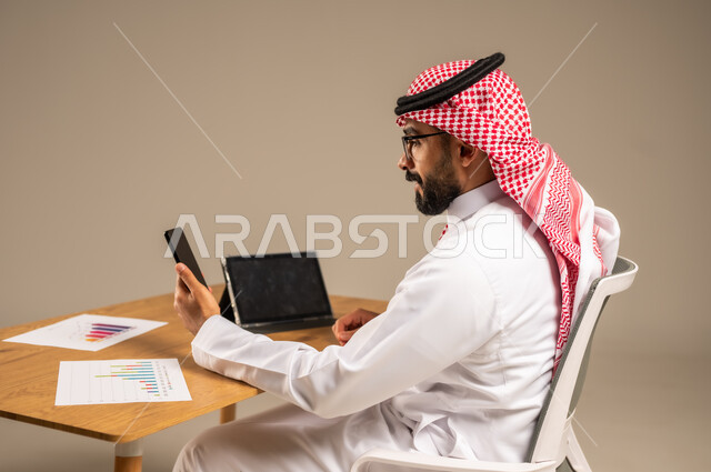 استخدام الهاتف المحمول في إدارة شؤون الأعمال عبر الانترنت، وظائف إدارية مكتبية أونلاين, دمج العمل مع التقنيات التكنولوجية، مفهوم العمل عن بعد، بورتريه من الخلف لرجل عربي خليجي سعودي يرتدي الثوب التقليدي والشماغ يجلس خلف المكتب وأمامه جهاز التابلت، خلفية رمادية