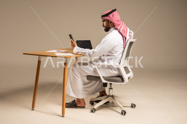 استخدام الهاتف المحمول في إدارة شؤون الأعمال عبر الانترنت، وظائف إدارية مكتبية أونلاين, دمج العمل مع التقنيات التكنولوجية، مفهوم العمل عن بعد، بورتريه من الخلف لرجل عربي خليجي سعودي يرتدي الثوب التقليدي والشماغ يجلس خلف المكتب وأمامه جهاز التابلت، خلفية رمادية