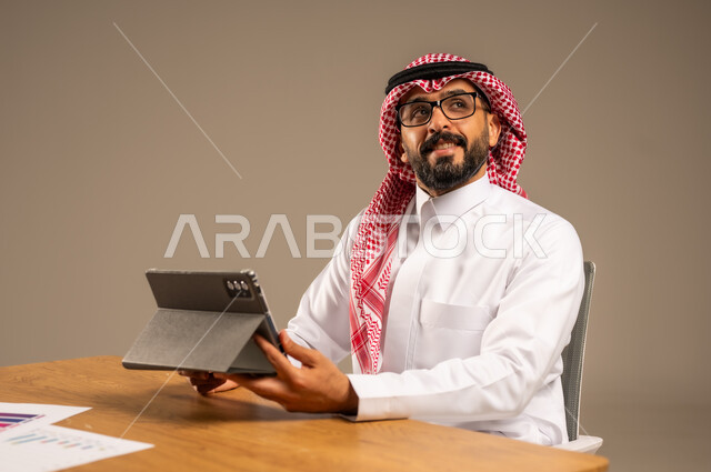 إدارة شؤون الأعمال عبر الانترنت، وظائف إدارية مكتبية أونلاين للشباب في السعودية, دمج العمل مع التقنيات التكنولوجية، مفهوم العمل عن بعد، بورتريه من الجانب لرجل عربي خليجي سعودي يرتدي الثوب التقليدي والشماغ يجلس خلف المكتب وأمامه جهاز التابلت، خلفية رمادية