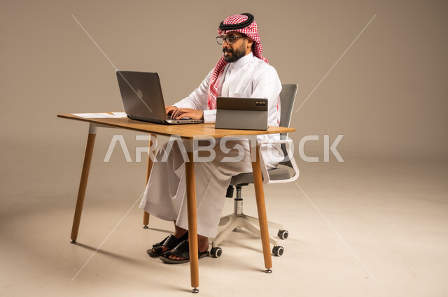 مهن ووظائف إدارية مكتبية أونلاين للشباب في السعودية, دمج العمل مع التقنيات التكنولوجية، مفهوم العمل عن بعد عبر الانترنت، بورتريه من الجانب لرجل عربي خليجي سعودي يرتدي الثوب التقليدي والشماغ يجلس خلف المكتب وأمامه الحاسوب المحمول وجهاز التابلت، خلفية رمادية