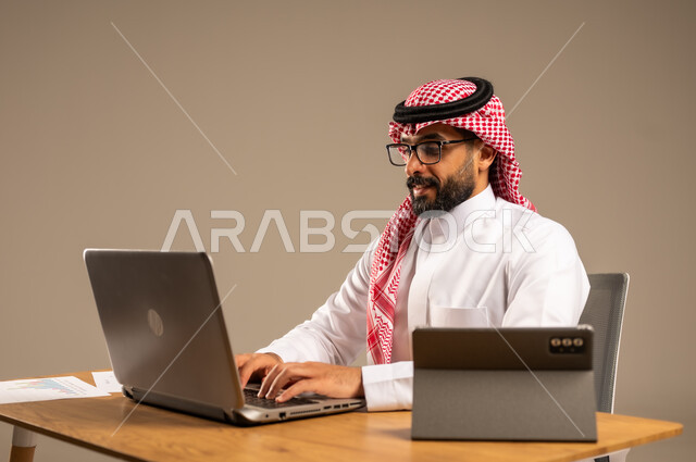 مهن ووظائف إدارية مكتبية أونلاين للشباب في السعودية, دمج العمل مع التقنيات التكنولوجية، مفهوم العمل عن بعد عبر الانترنت، بورتريه من الجانب لرجل عربي خليجي سعودي يرتدي الثوب التقليدي والشماغ يجلس خلف المكتب وأمامه الحاسوب المحمول وجهاز التابلت، خلفية رمادية