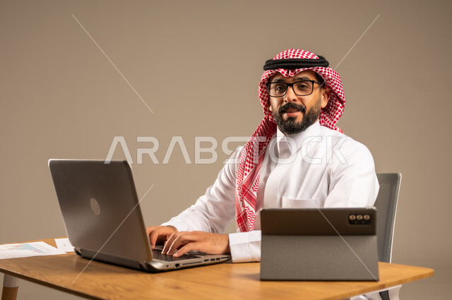 مهن ووظائف إدارية مكتبية أونلاين للشباب في السعودية, دمج العمل مع التقنيات التكنولوجية، مفهوم العمل عن بعد عبر الانترنت، بورتريه من الجانب لرجل عربي خليجي سعودي يرتدي الثوب التقليدي والشماغ يجلس خلف المكتب وأمامه الحاسوب المحمول وجهاز التابلت، خلفية رمادية