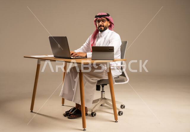 مهن ووظائف إدارية مكتبية أونلاين للشباب في السعودية, دمج العمل مع التقنيات التكنولوجية، مفهوم العمل عن بعد عبر الانترنت، بورتريه من الجانب لرجل عربي خليجي سعودي يرتدي الثوب التقليدي والشماغ يجلس خلف المكتب وأمامه الحاسوب المحمول وجهاز التابلت، خلفية رمادية