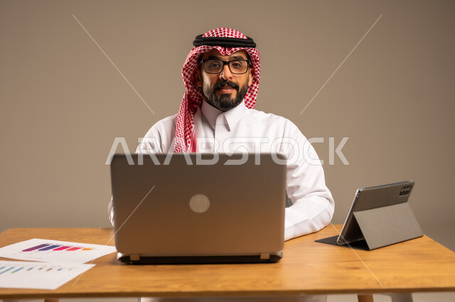 The concept of remote work via the Internet, integrating work with technology and modern technology, online office administrative jobs and professions for young people in Saudi Arabia, portrait of a Saudi Arabian Gulf man wearing traditional thobe and shemagh sitting behind a desk with a laptop and tablet in front of him, gray background