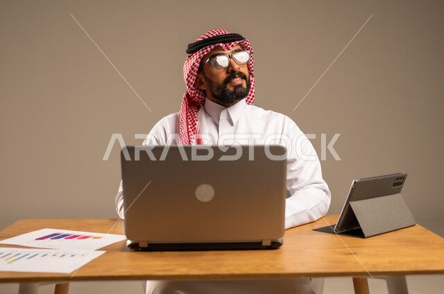 The concept of remote work via the Internet, integrating work with technology and modern technology, online office administrative jobs and professions for young people in Saudi Arabia, portrait of a Saudi Arabian Gulf man wearing traditional thobe and shemagh sitting behind a desk with a laptop and tablet in front of him, gray background