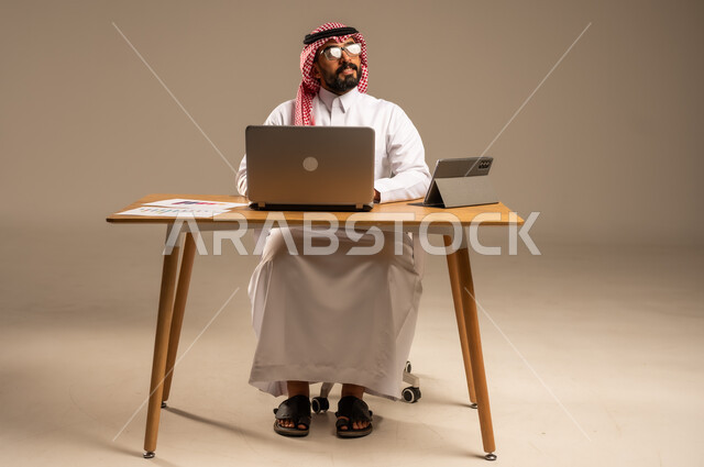 The concept of remote work via the Internet, integrating work with technology and modern technology, online office administrative jobs and professions for young people in Saudi Arabia, portrait of a Saudi Arabian Gulf man wearing traditional thobe and shemagh sitting behind a desk with a laptop and tablet in front of him, gray background