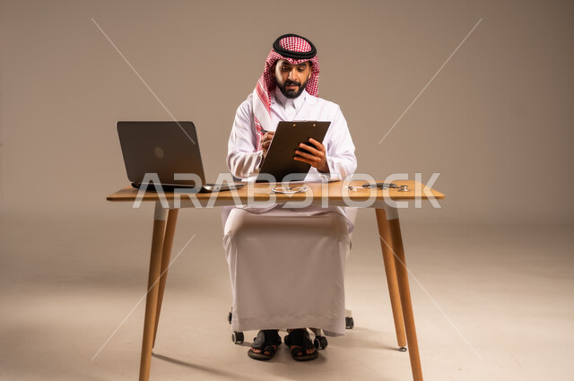 Providing services to patients, modifying and recording notes in their files, using technology in health research and development, conducting remote medical consultations, portrait of a young Saudi Arabian Gulf man wearing a medical coat and a shemagh sitting behind a desk with a laptop and an examination stethoscope in front of him, gray background